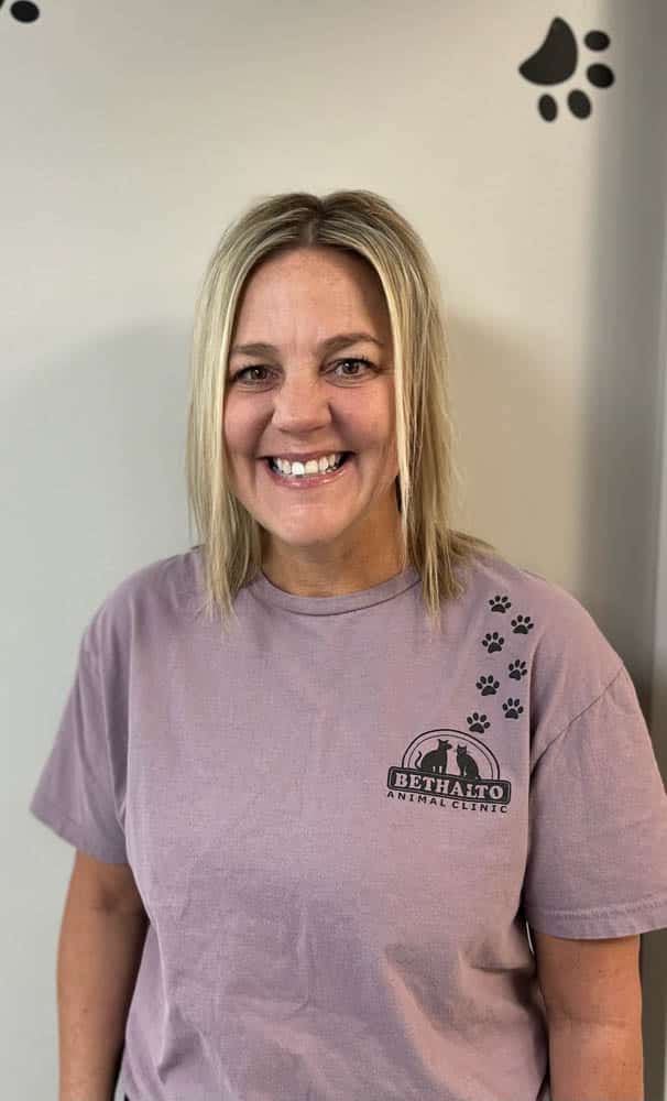 Smiling woman with blonde hair wears a purple Bethalto Animal Clinic t-shirt, standing in front of a light wall with black paw prints.