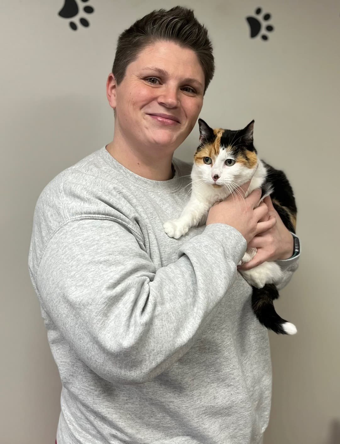 A person wearing a gray sweatshirt holds a calico cat in their arms, standing in front of a wall decorated with black paw prints.