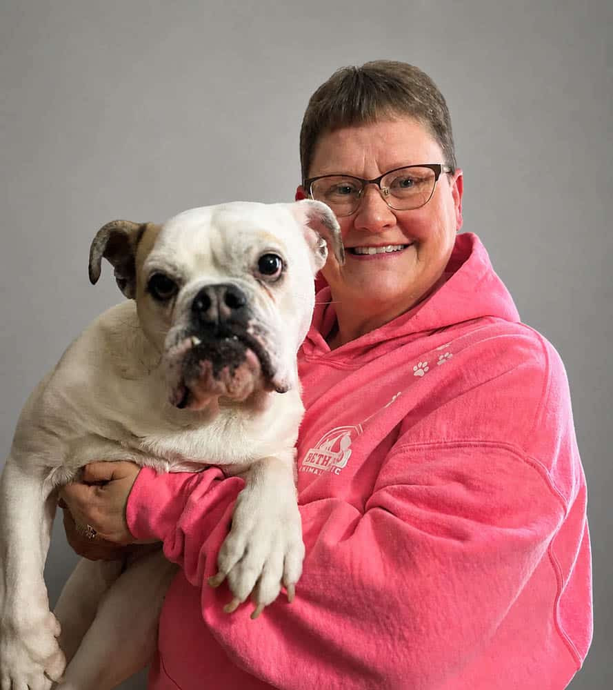 A person wearing glasses and a pink hoodie is smiling and holding a white and brown bulldog against a plain gray background.
